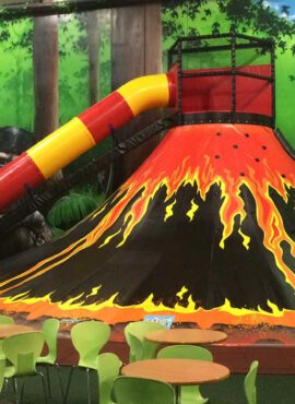 Indoor play area featuring a vibrant volcano-themed slide with a bright yellow and red tube, surrounded by green seating and a forest backdrop, ideal for children's entertainment and adventure.