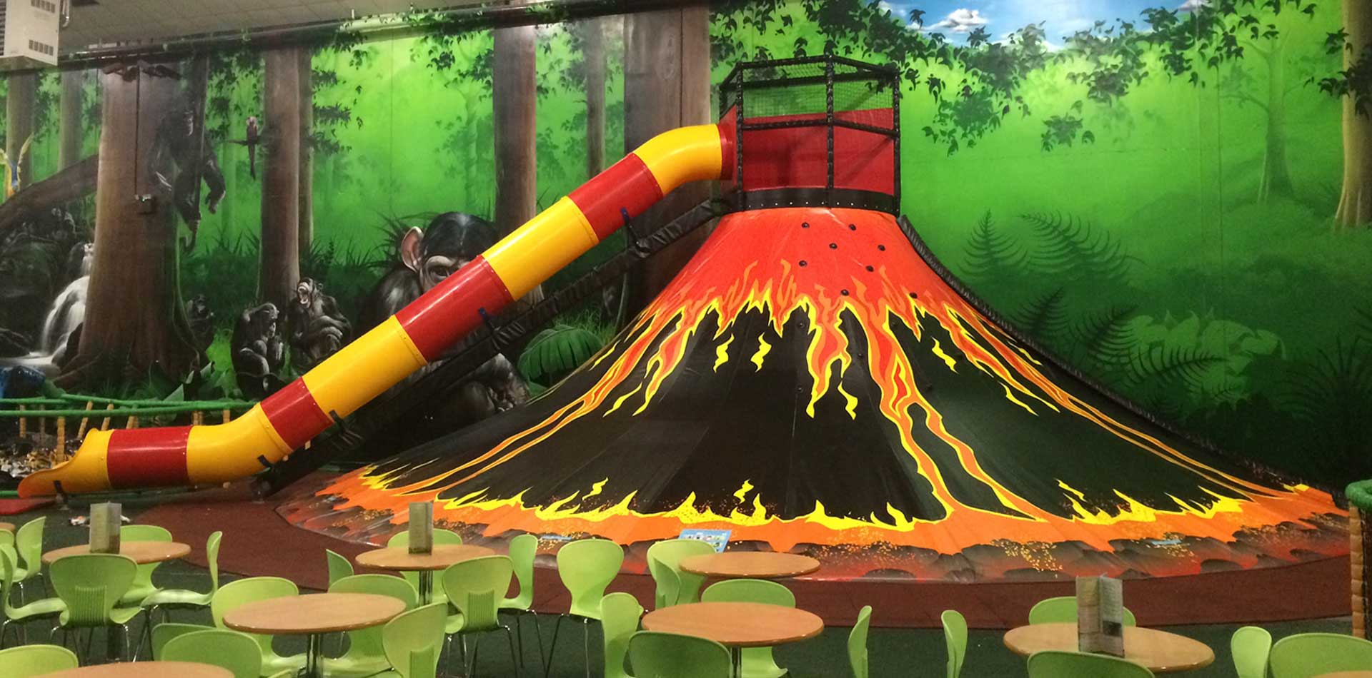 Indoor play area featuring a vibrant volcano-themed slide with a bright yellow and red tube, surrounded by green seating and a forest backdrop, ideal for children's entertainment and adventure.