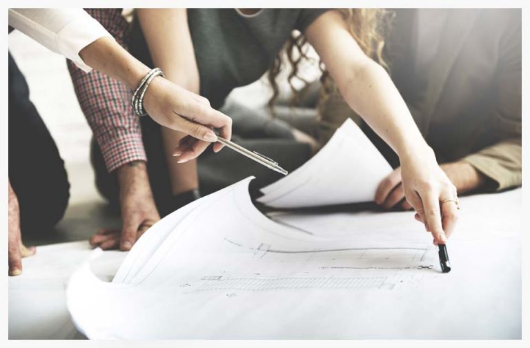 A group of professionals collaboratively reviewing architectural blueprints on a table, with hands pointing and holding pens, illustrating teamwork in project planning.