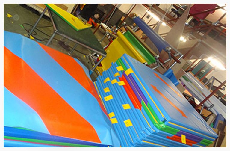 Colourful gym mats stacked in a sports facility, featuring a variety of shapes and sizes in blue, orange, and green, designed for safe landing and cushioning during physical activities.