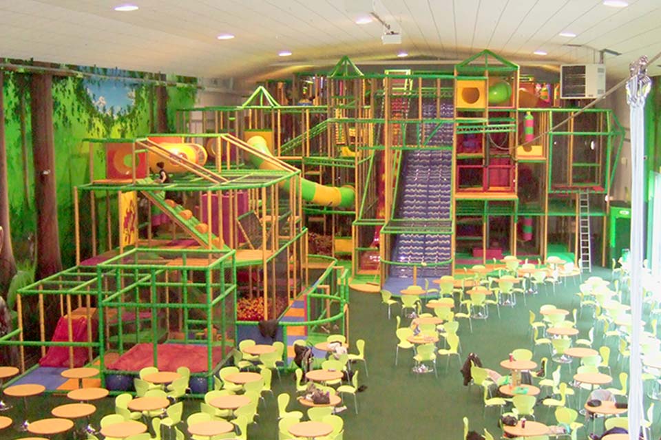 Indoor play area featuring a large, multi-level climbing frame with slides, tunnels, and soft play equipment, surrounded by brightly coloured tables and chairs for children.