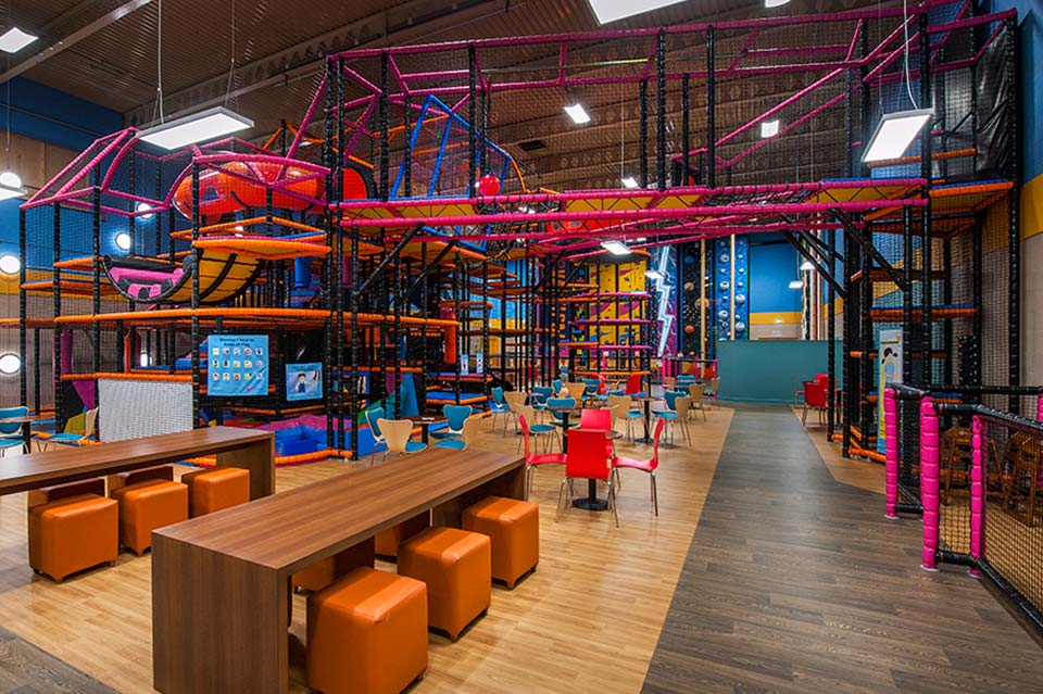 Indoor play area featuring a vibrant climbing structure with orange and pink elements, surrounded by seating areas with colourful chairs and tables, designed for children's entertainment and activities.