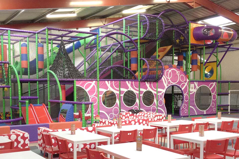 Indoor children's play area featuring colourful climbing structures, slides, and soft play equipment, with vibrant pink and green designs and red dining tables.
