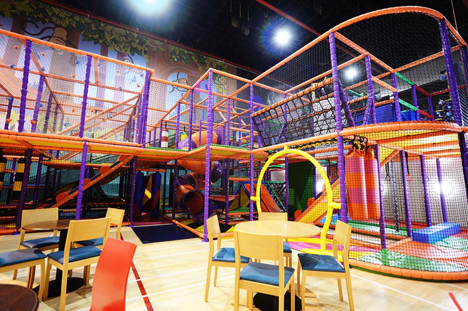 Indoor play area featuring a vibrant multi-level climbing frame with slides, tunnels, and various play equipment, surrounded by seating for parents and guardians. Ideal for children's entertainment and physical activity.