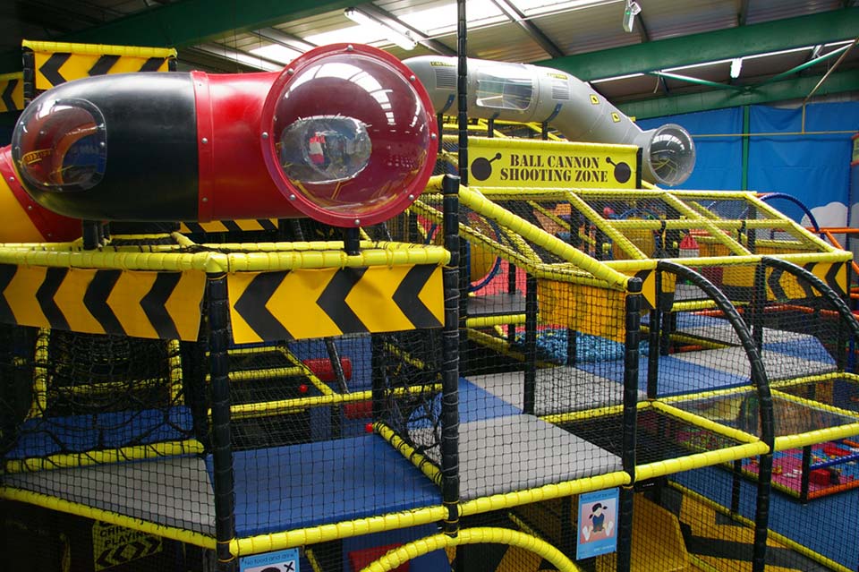 Indoor soft play area featuring bright yellow and red climbing structures, slides, and tunnels designed for children's entertainment and activity. Safety mesh and colourful barriers enhance the play environment.