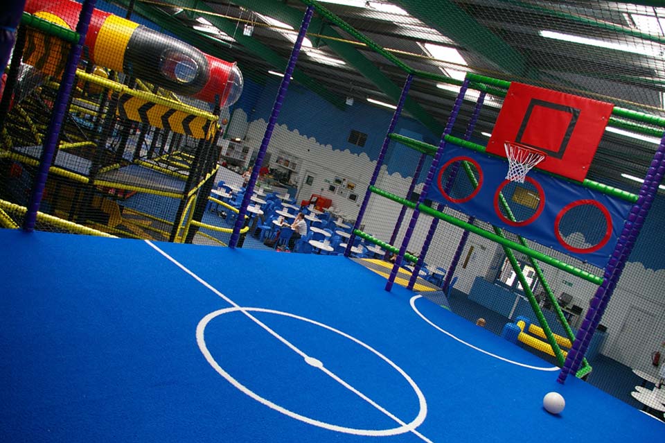 Indoor play area featuring colourful climbing structures, trampolines, and a basketball hoop on a vibrant blue floor, designed for children's active play and exercise.