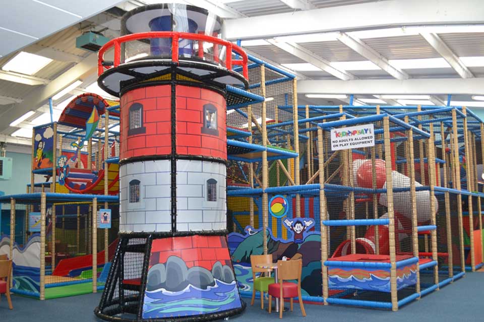 Colourful indoor play area featuring a large red and white lighthouse structure, surrounded by climbing frames and soft play equipment, designed for children's entertainment and physical activity.