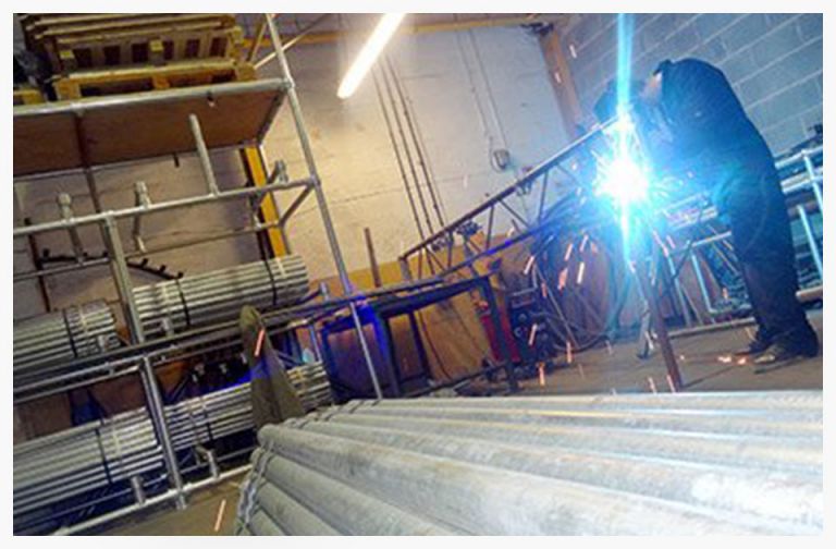 Welder working on metal scaffolding in a workshop, with sparks flying from the welding process and various metal pipes stacked in the foreground.
